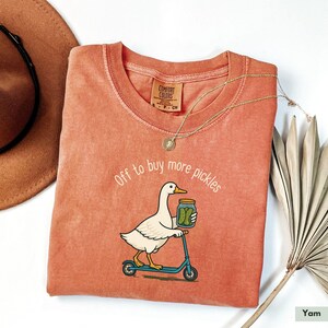 May include: A coral-coloured Comfort Colors t-shirt with the phrase "Off to buy more pickles" and a graphic of a duck riding a scooter while holding a jar of pickles. A gold necklace and a brown hat are also visible.