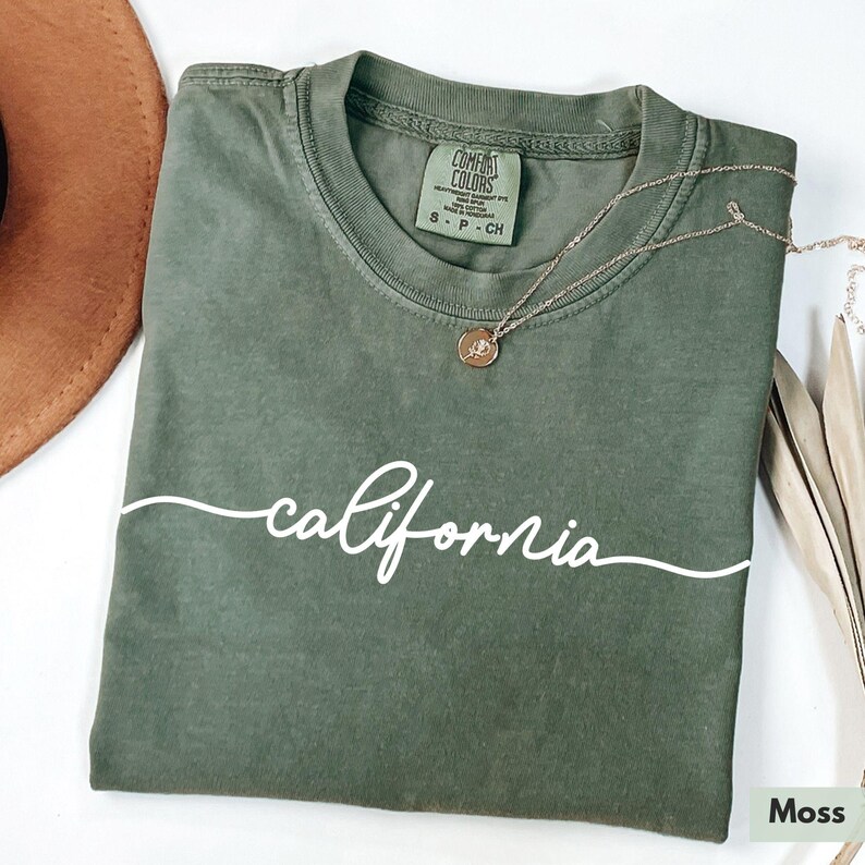 May include: A moss green t-shirt with the word "california" written in white cursive script. The shirt has a tag that reads "Comfort Colors" and "S - P - CH".
