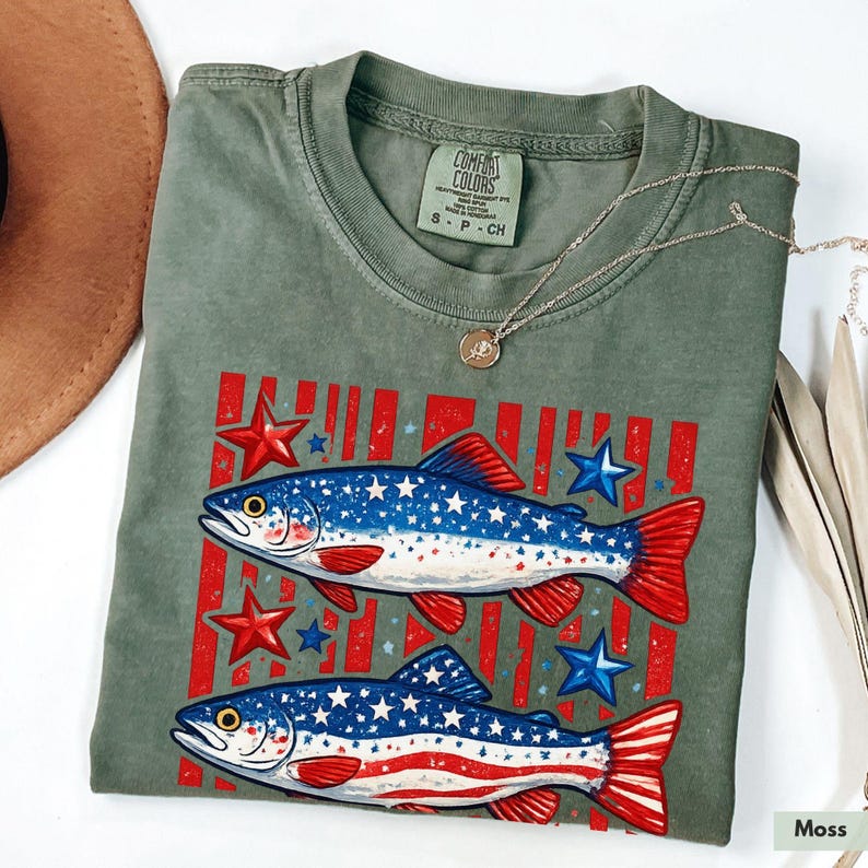 May include: A moss green Comfort Colors t-shirt featuring a patriotic design. Two fish are depicted with blue and white star patterns and red and white stripes. Red stars and blue stars are scattered around the fish. A silver necklace is draped on the shirt.