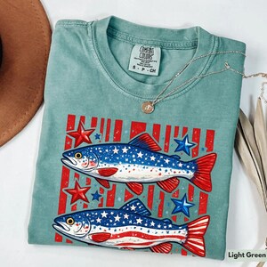 May include: Light green Comfort Colors t-shirt featuring two fish with red and blue accents, stars, and stripes. The fish have a patriotic design with stars and stripes. A silver necklace with a pendant is on top of the shirt.