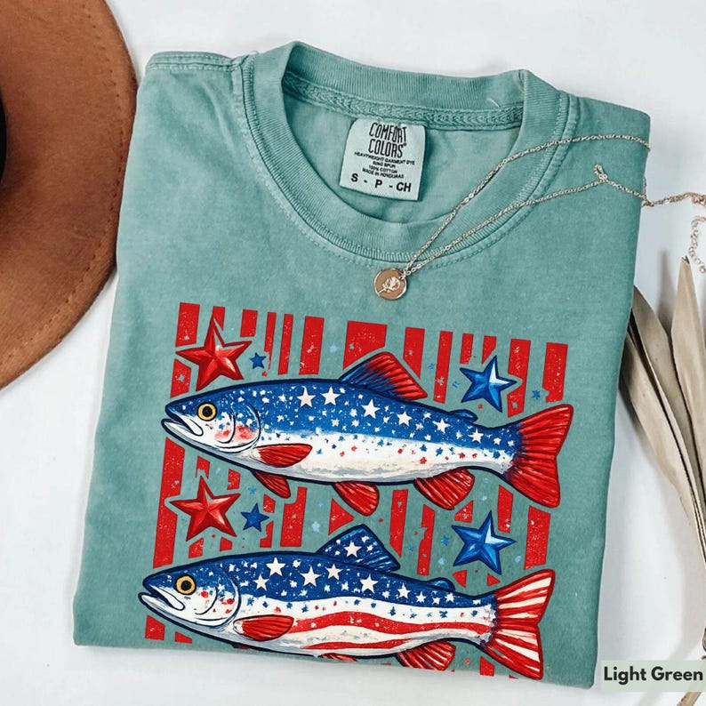 May include: Light green Comfort Colors t-shirt featuring two fish with red and blue accents, stars, and stripes. The fish have a patriotic design with stars and stripes. A silver necklace with a pendant is on top of the shirt.