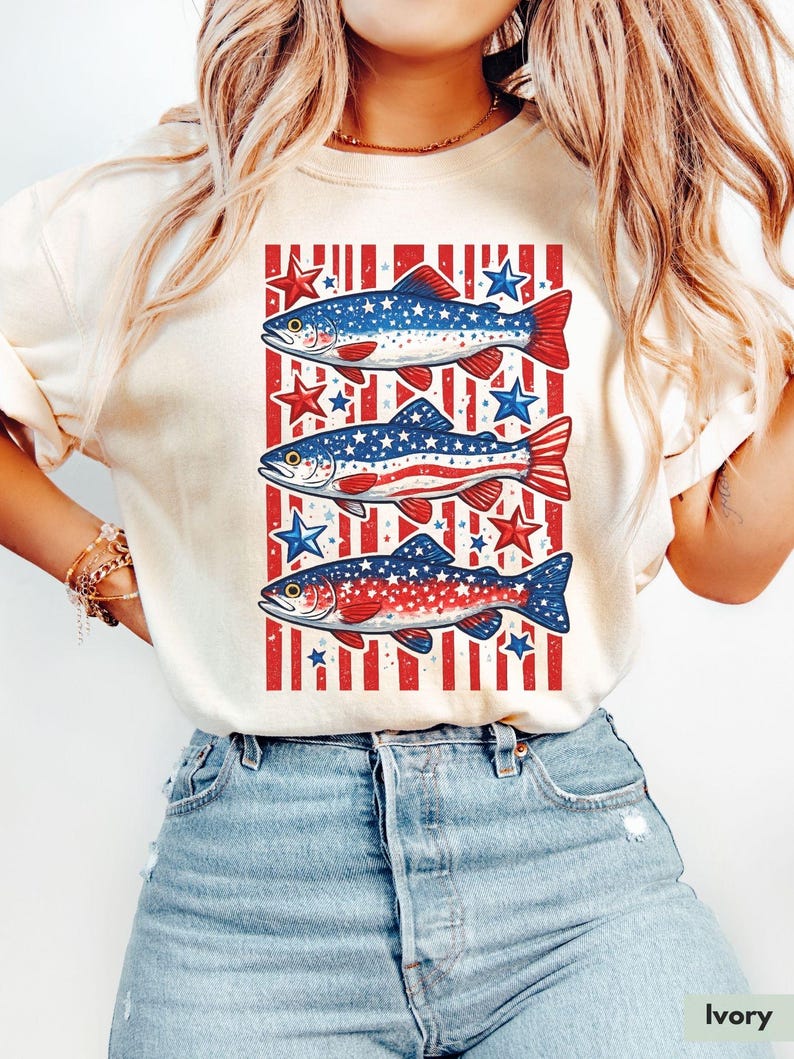 May include: Ivory-colored t-shirt featuring a patriotic design of three fish with red and blue patterns, set against a red and white striped background. The fish are adorned with stars, creating a festive and eye-catching graphic.