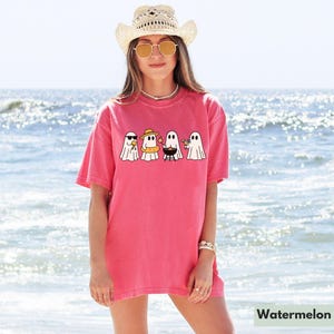 May include: A watermelon-colored t-shirt featuring a graphic of four cartoon ghosts enjoying summer activities. One ghost wears sunglasses, another a sun hat and inflatable ring, a third is grilling, and the last holds a drink. The word "Watermelon" is at the bottom.