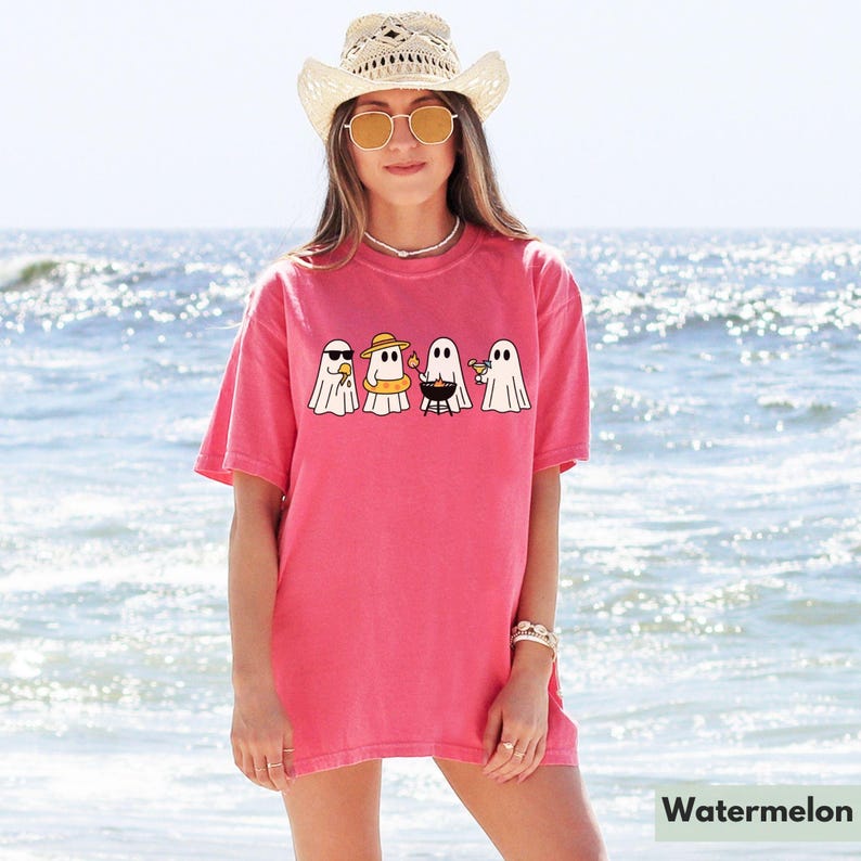 May include: A watermelon-colored t-shirt featuring a graphic of four cartoon ghosts enjoying summer activities. One ghost wears sunglasses, another a sun hat and inflatable ring, a third is grilling, and the last holds a drink. The word "Watermelon" is at the bottom.