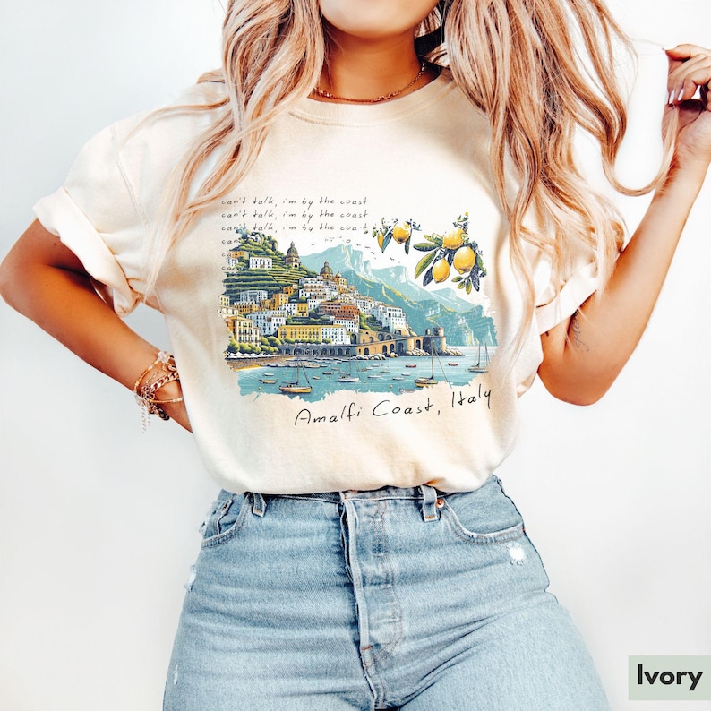 以下が含まれることがあります： レモンの木と「can't talk, i'm by the coast」と「Amalfi Coast, Italy」のテキストが書かれた、イタリアのアマルフィ海岸のグラフィックがプリントされた白いTシャツ。