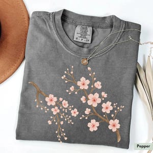 May include: A gray t-shirt with a pink cherry blossom design. The shirt has a tag that says "Comfort Colors" and "S - P - CH".