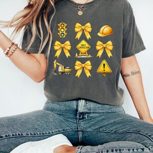 May include: A gray t-shirt with yellow construction-themed graphics, including a traffic light, hard hat, cones, a construction sign that says "Under Construction", and a backhoe. The graphics are surrounded by yellow bows.