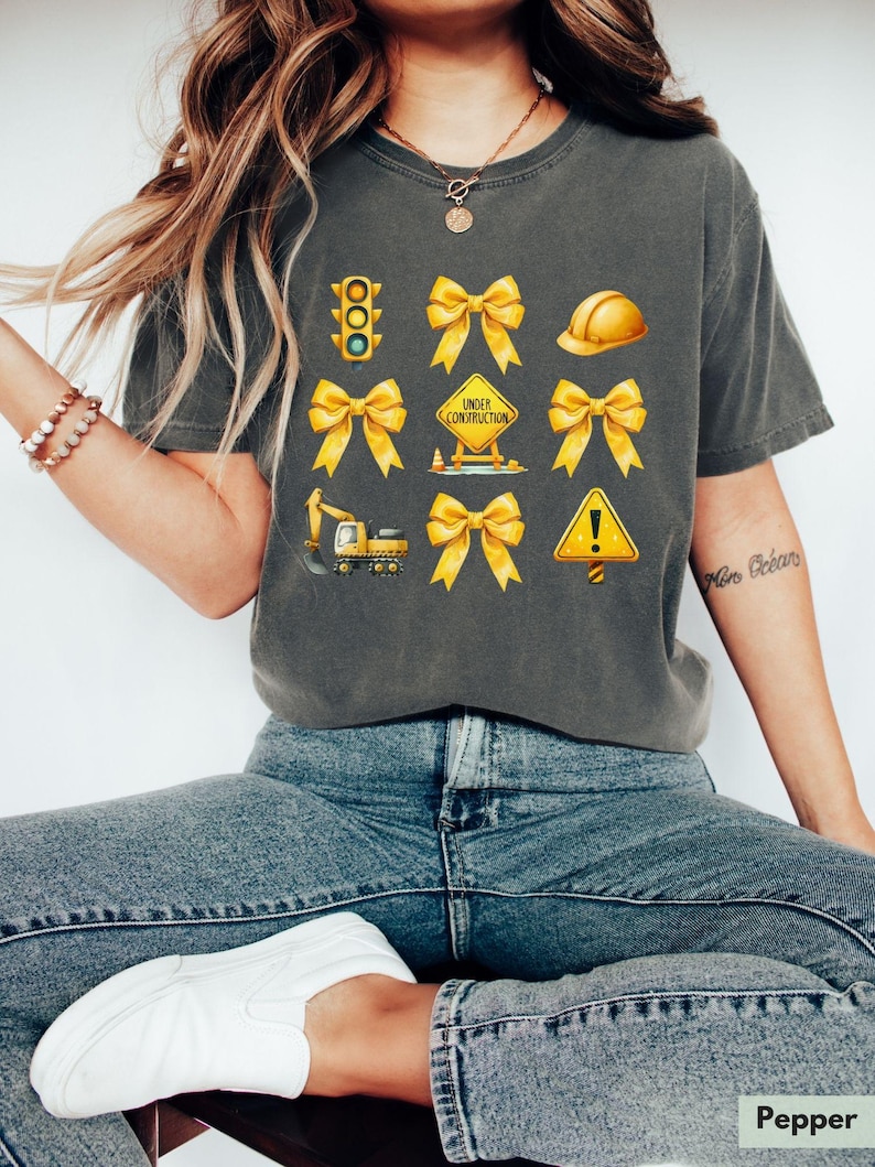 May include: A gray t-shirt with yellow construction-themed graphics, including a traffic light, hard hat, cones, a construction sign that says "Under Construction", and a backhoe. The graphics are surrounded by yellow bows.