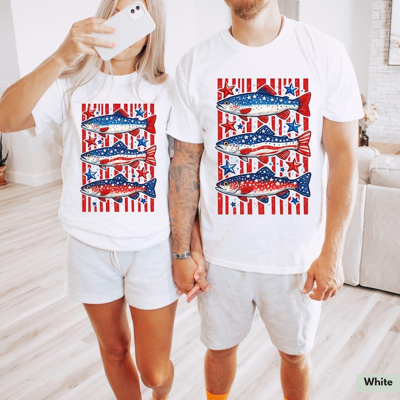May include: White t-shirts with a patriotic fish design. Each shirt features three fish with red, white, and blue patterns, set against a background of red stripes and blue stars. The shirts are worn by two people.