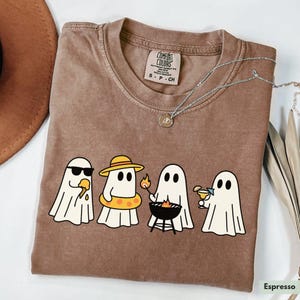 May include: Espresso-colored t-shirt featuring a graphic of four cartoon ghosts enjoying summer activities. One ghost wears sunglasses and eats ice cream, another wears a hat and floatie, one grills, and the last sips a cocktail.