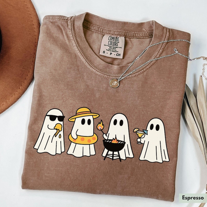 May include: Espresso-colored t-shirt featuring a graphic of four cartoon ghosts enjoying summer activities. One ghost wears sunglasses and eats ice cream, another wears a hat and floatie, one grills, and the last sips a cocktail.