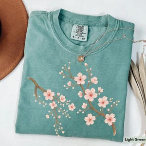 May include: Light green t-shirt with a pink cherry blossom design. The shirt has a crew neck and a tag that reads "Comfort Colors" with the size "S - P - CH".
