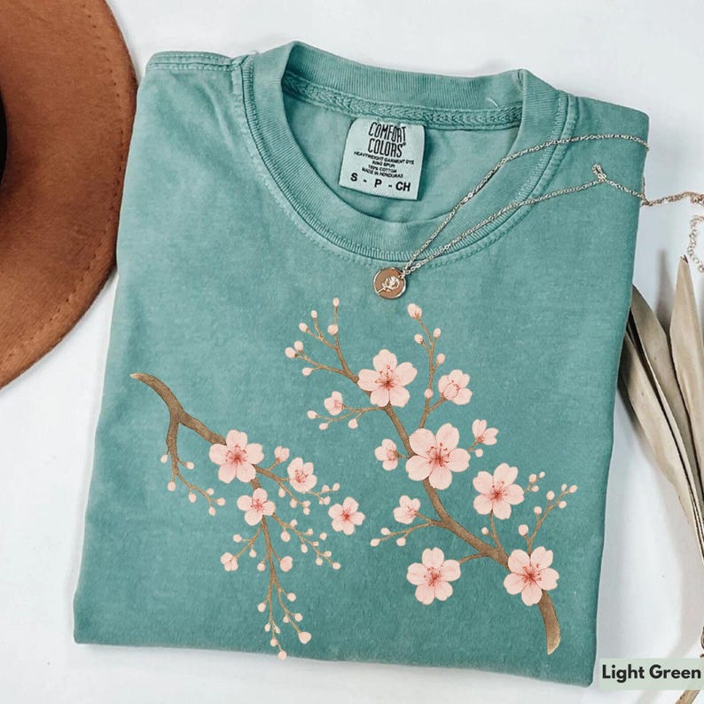 May include: Light green t-shirt with a pink cherry blossom design. The shirt has a crew neck and a tag that reads "Comfort Colors" with the size "S - P - CH".