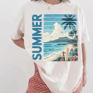 May include: Ivory-colored t-shirt featuring a tropical beach scene with the word "SUMMER" in blue vertical text. The graphic includes palm trees, surfboards, and ocean waves. The shirt is paired with red and white checkered shorts.