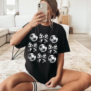 Puede incluir: Camiseta negra con un gr&aacute;fico de balones de f&uacute;tbol y lazos. Los balones de f&uacute;tbol son blancos con costuras negras y los lazos son de cuadros negros y blancos.
