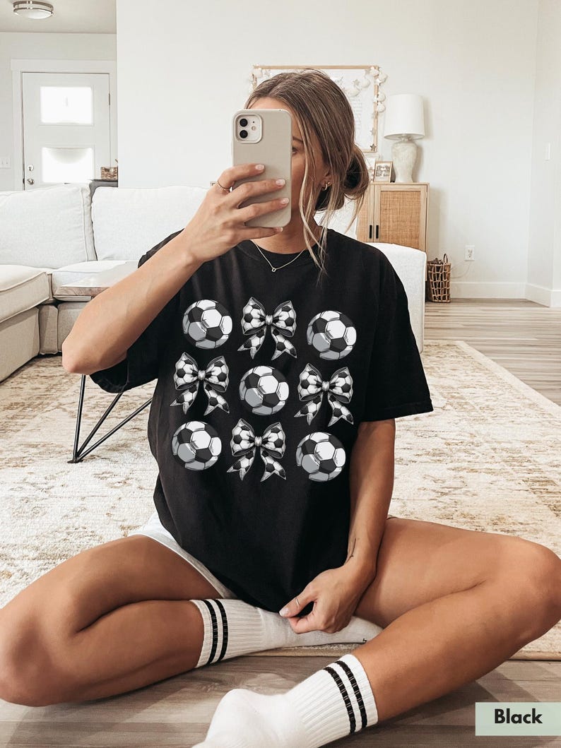 Puede incluir: Camiseta negra con un gr&aacute;fico de balones de f&uacute;tbol y lazos. Los balones de f&uacute;tbol son blancos con costuras negras y los lazos son de cuadros negros y blancos.