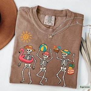 May include: A brown t-shirt with a faded look features a graphic of three skeletons wearing summer accessories. The skeletons are holding a beach ball, a tropical drink, and a flamingo float. The text "COMFORT COLORS" is visible on a tag attached to the shirt.