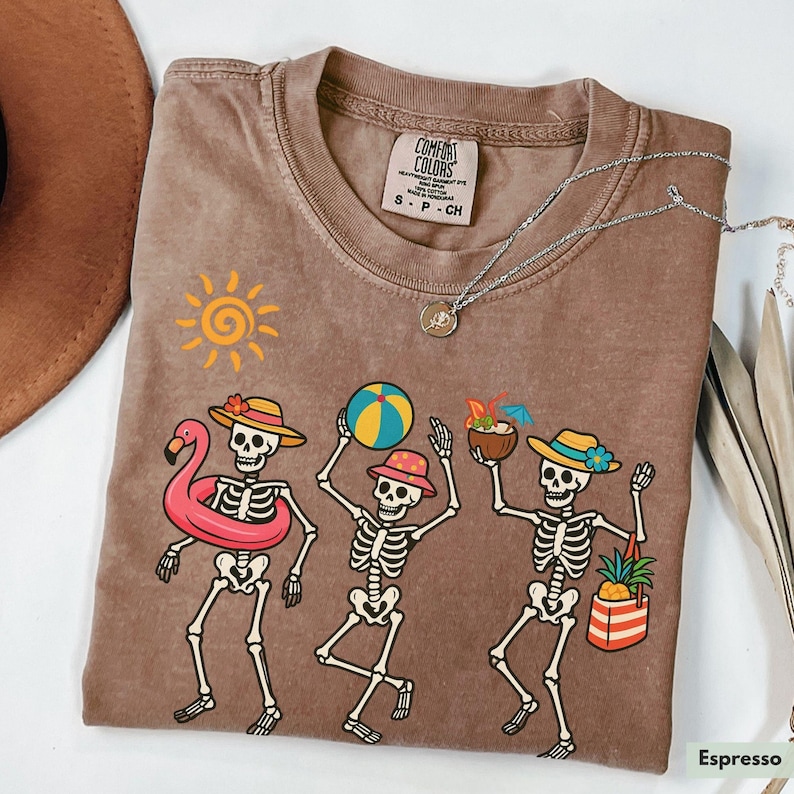 May include: A brown t-shirt with a faded look features a graphic of three skeletons wearing summer accessories. The skeletons are holding a beach ball, a tropical drink, and a flamingo float. The text "COMFORT COLORS" is visible on a tag attached to the shirt.