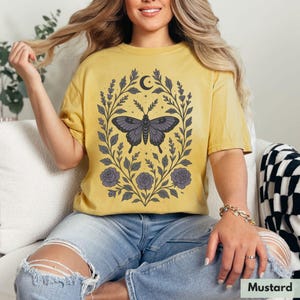 May include: A mustard yellow t-shirt with a floral design featuring a butterfly and a crescent moon. The design is in shades of purple and gray.