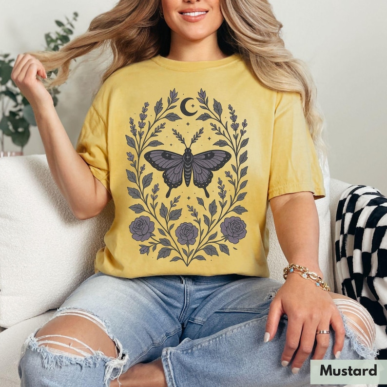 May include: A mustard yellow t-shirt with a floral design featuring a butterfly and a crescent moon. The design is in shades of purple and gray.