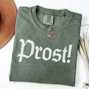 Puede incluir: Una camiseta verde con la palabra "Prost!" impresa en blanco en la parte delantera. La camiseta est&aacute; hecha de un material suave y c&oacute;modo y tiene un aspecto vintage.
