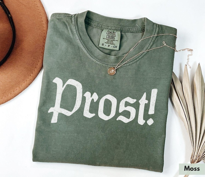 Puede incluir: Una camiseta verde con la palabra "Prost!" impresa en blanco en la parte delantera. La camiseta est&aacute; hecha de un material suave y c&oacute;modo y tiene un aspecto vintage.