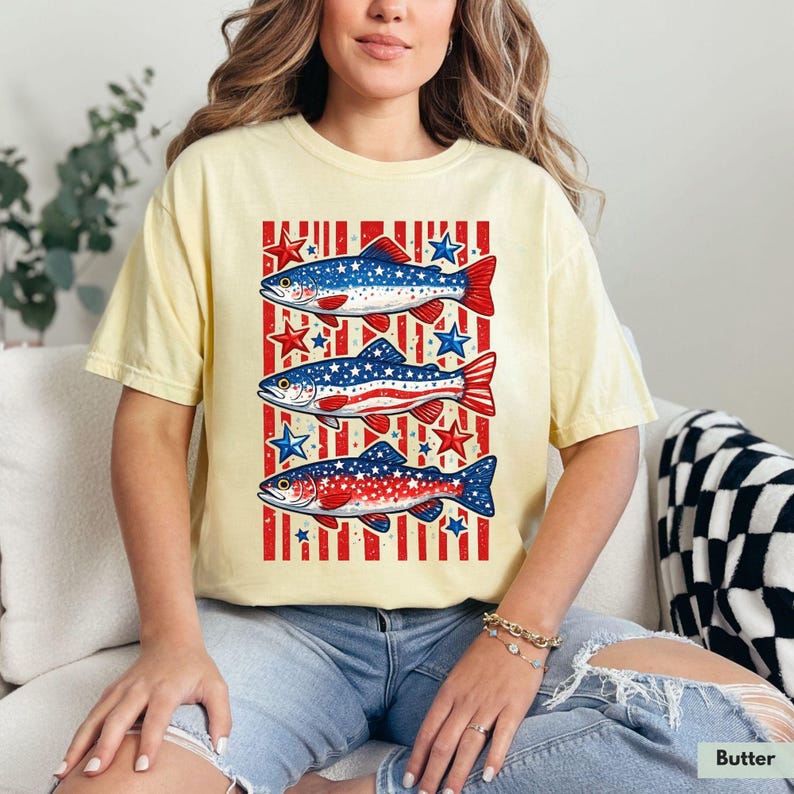 May include: Butter-colored t-shirt featuring a patriotic design of three fish with red, white, and blue patterns. The fish are set against a background of red stripes and blue stars. The shirt is a relaxed fit.