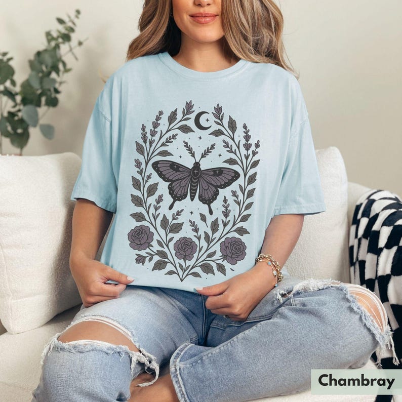 May include: A light blue t-shirt with a floral design featuring a butterfly and a crescent moon. The design is in shades of gray and black.