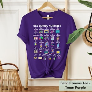 May include: Purple t-shirt with a graphic of an alphabet with old school items. The graphic says "Old School Alphabet" and each letter of the alphabet is paired with a word and an image. For example, "Aa" is paired with "Arcade" and an image of an arcade game. The shirt is a Bella Canvas tee.