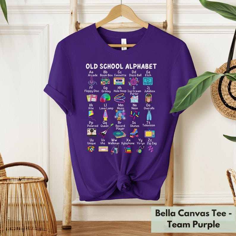 May include: Purple t-shirt with a graphic of an alphabet with old school items. The graphic says "Old School Alphabet" and each letter of the alphabet is paired with a word and an image. For example, "Aa" is paired with "Arcade" and an image of an arcade game. The shirt is a Bella Canvas tee.