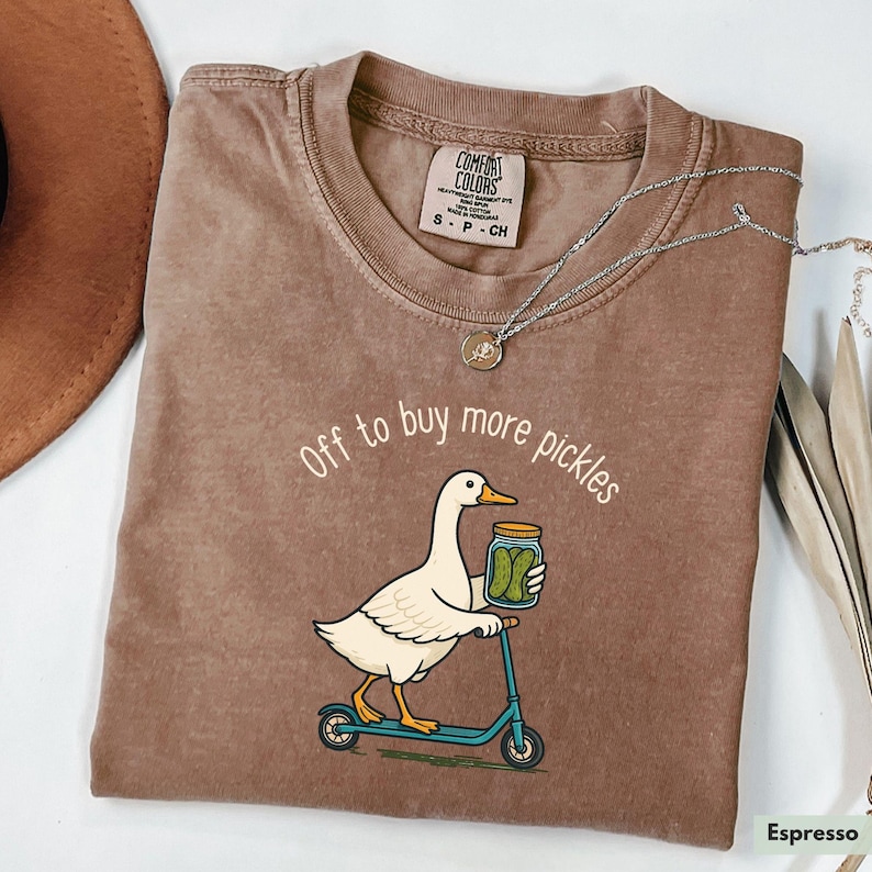 May include: Espresso-coloured t-shirt featuring a white duck on a blue scooter, carrying a jar of pickles. The text reads "Off to buy more pickles". The shirt has a Comfort Colors label and is a small size.