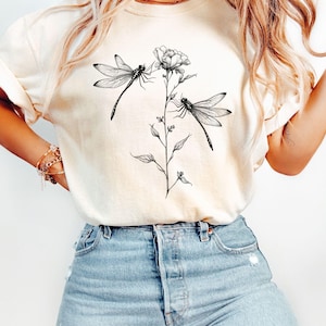 May include: A white t-shirt with a black and white graphic of two dragonflies on a flower stem. The shirt is being worn by a person with long blonde hair and blue jeans.