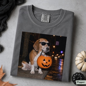 Könnte beinhalten: Ein anthrazitfarbenes T-Shirt mit einem Grafikdruck eines Beagle-Hundes, der eine schwarze Sonnenbrille trägt und einen orangefarbenen Kunststoff-Kürbis-Süßigkeiteneimer im Maul hält. Das Shirt hat ein Comfort Colors-Etikett am inneren Halsausschnitt.