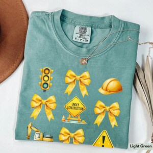 May include: A light green t-shirt with yellow construction-themed graphics. The graphics include a traffic light, a construction sign that says "Under Construction", a hard hat, and several yellow bows. The shirt also has a small tag that says "Comfort Colors" and "S-P-CH".