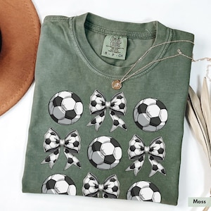 Puede incluir: Una camiseta verde con un patr&oacute;n de bal&oacute;n de f&uacute;tbol y lazo. La camiseta tiene el texto "Comfort Colors" en la etiqueta.