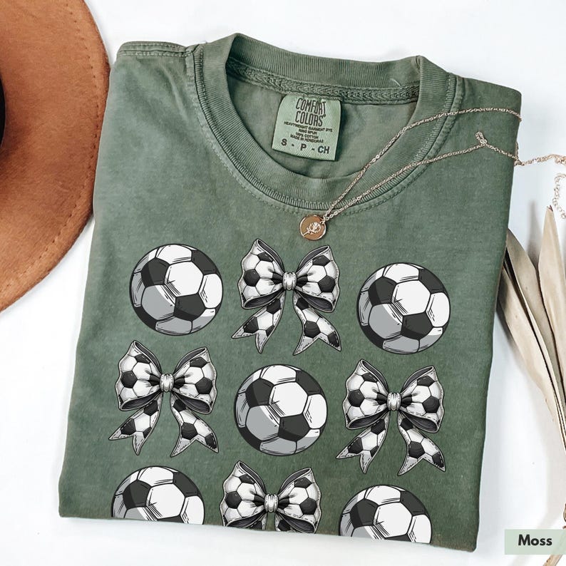 Puede incluir: Una camiseta verde con un patr&oacute;n de bal&oacute;n de f&uacute;tbol y lazo. La camiseta tiene el texto "Comfort Colors" en la etiqueta.