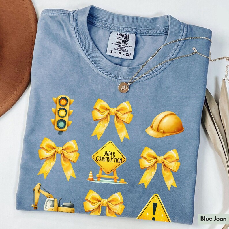 May include: A blue jean t-shirt with yellow construction-themed graphics. The graphics include a traffic light, a hard hat, a construction sign that says "Under Construction", and several yellow bows. The shirt has a tag that says "Comfort Colors".