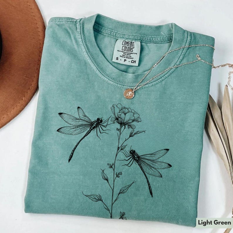 May include: Light green t-shirt with a black dragonfly and flower design. The shirt has the text "Comfort Colors" and "S - P - CH" printed on the front.