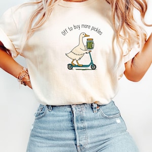 May include: Ivory-coloured t-shirt featuring a duck graphic on a scooter, holding a pickle jar, with the text "Off to buy more pickles". The model is wearing blue jeans.