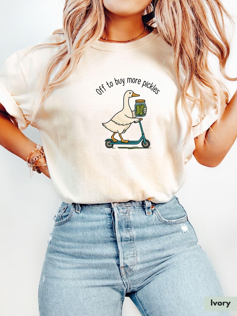 May include: Ivory-coloured t-shirt featuring a duck graphic on a scooter, holding a pickle jar, with the text "Off to buy more pickles". The model is wearing blue jeans.
