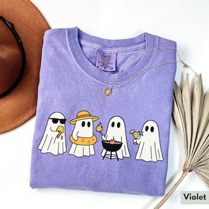 May include: A violet Comfort Colors t-shirt featuring a graphic of four cartoon ghosts enjoying summer activities. One ghost wears sunglasses and eats ice cream, another wears a hat and floatie, a third grills, and the last one sips a cocktail.