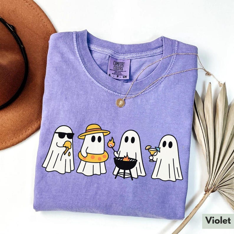 May include: A violet Comfort Colors t-shirt featuring a graphic of four cartoon ghosts enjoying summer activities. One ghost wears sunglasses and eats ice cream, another wears a hat and floatie, a third grills, and the last one sips a cocktail.