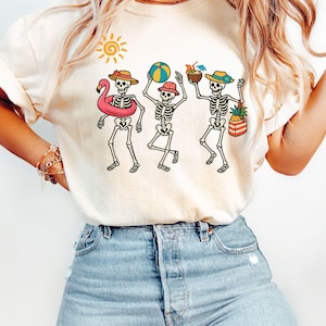May include: A white t-shirt with a graphic of three skeletons wearing summer accessories, including a flamingo float, a beach ball, and a pineapple. The skeletons are wearing hats and sunglasses. The shirt is on a model wearing blue jeans.