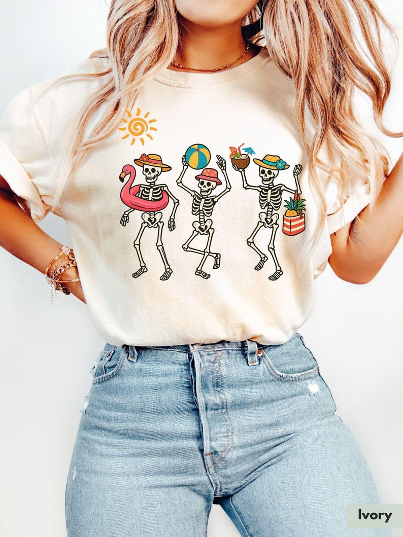 May include: A white t-shirt with a graphic of three skeletons wearing summer accessories, including a flamingo float, a beach ball, and a pineapple. The skeletons are wearing hats and sunglasses. The shirt is on a model wearing blue jeans.