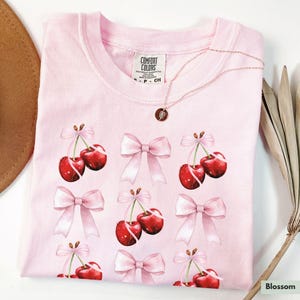 May include: Pink t-shirt with a repeating pattern of pink bows and red cherries. The shirt has the text "Comfort Colors" on the tag.