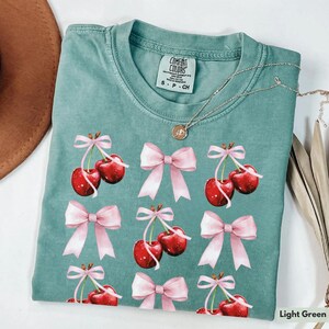 May include: A light green t-shirt with a watercolor print of pink bows and red cherries. The shirt has the text "Comfort Colors" and "S - P - CH" printed on the front.