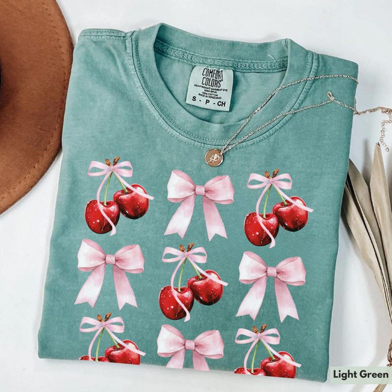 May include: A light green t-shirt with a watercolor print of pink bows and red cherries. The shirt has the text "Comfort Colors" and "S - P - CH" printed on the front.