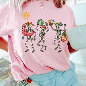 May include: Pink t-shirt with a graphic of three skeletons on vacation. The skeletons are wearing hats, sunglasses, and holding beach items like a beach ball, a pineapple, and a flamingo float.