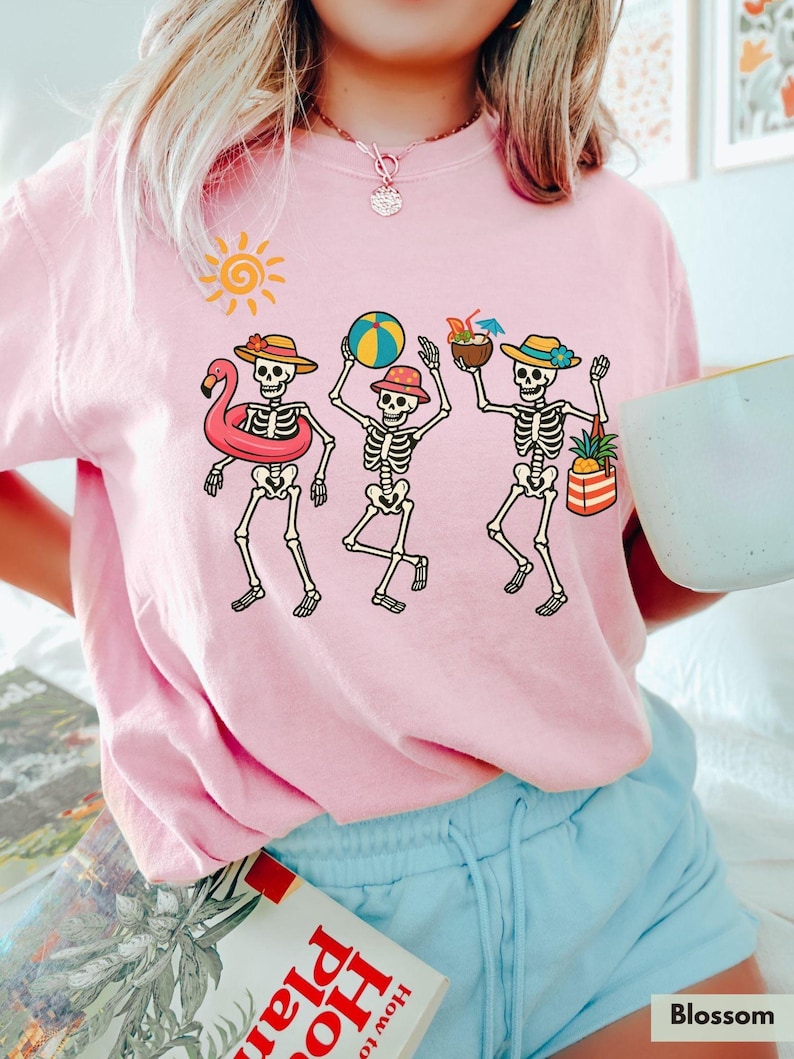 May include: Pink t-shirt with a graphic of three skeletons on vacation. The skeletons are wearing hats, sunglasses, and holding beach items like a beach ball, a pineapple, and a flamingo float.