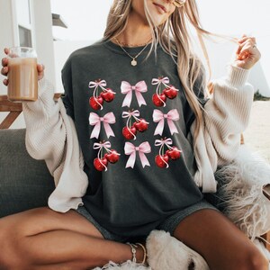 May include: A dark gray t-shirt with a graphic of red cherries with pink bows. The cherries are arranged in a grid pattern.